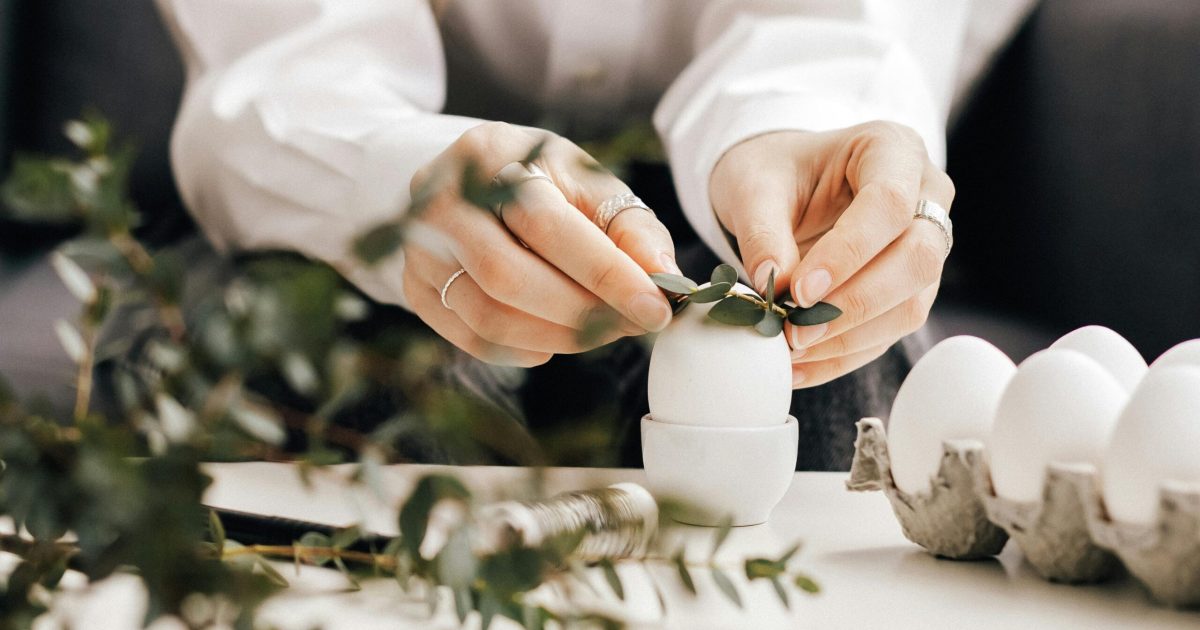 pexels-photo-6897679-6897679 Hands arranging leaves on white eggs for an Easter decoration indoors, creating a natural festive look.